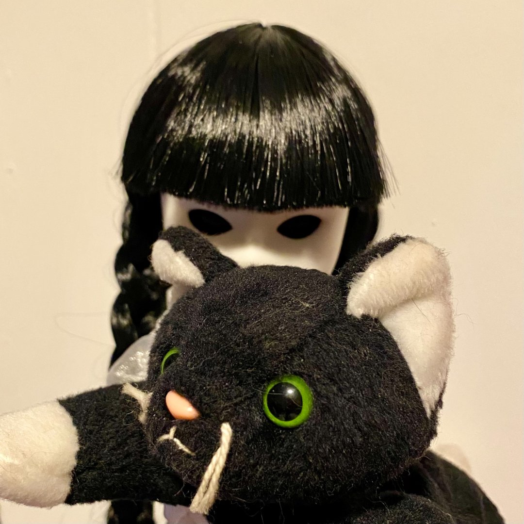 An eerie doll with black hair and hollow eyes holds a black plush cat with green eyes. The background is minimalist and light in color.