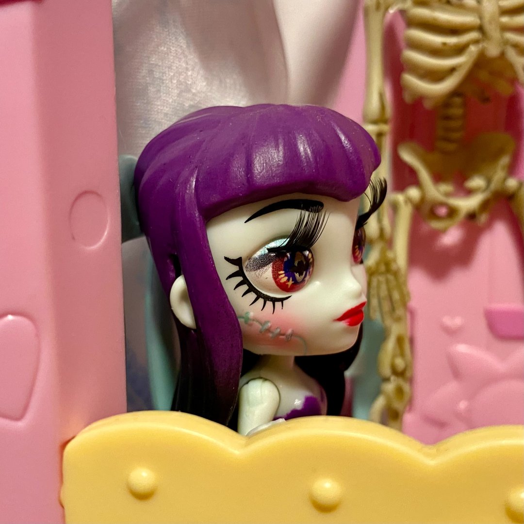 A close-up of a doll with purple hair and striking makeup features. The doll is leaning against a pink and yellow structure, with a skeletal decoration visible in the background.