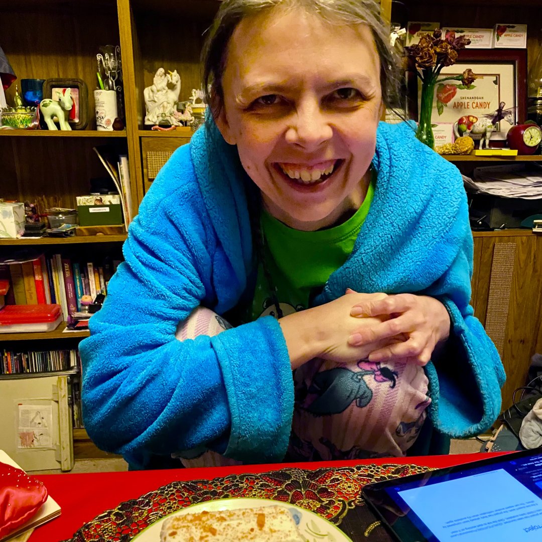 A smiling person wearing a blue robe and green shirt is sitting at a table with a plate of food in front of them. The background features bookshelves and various decorative items. The setting appears cozy and personal.