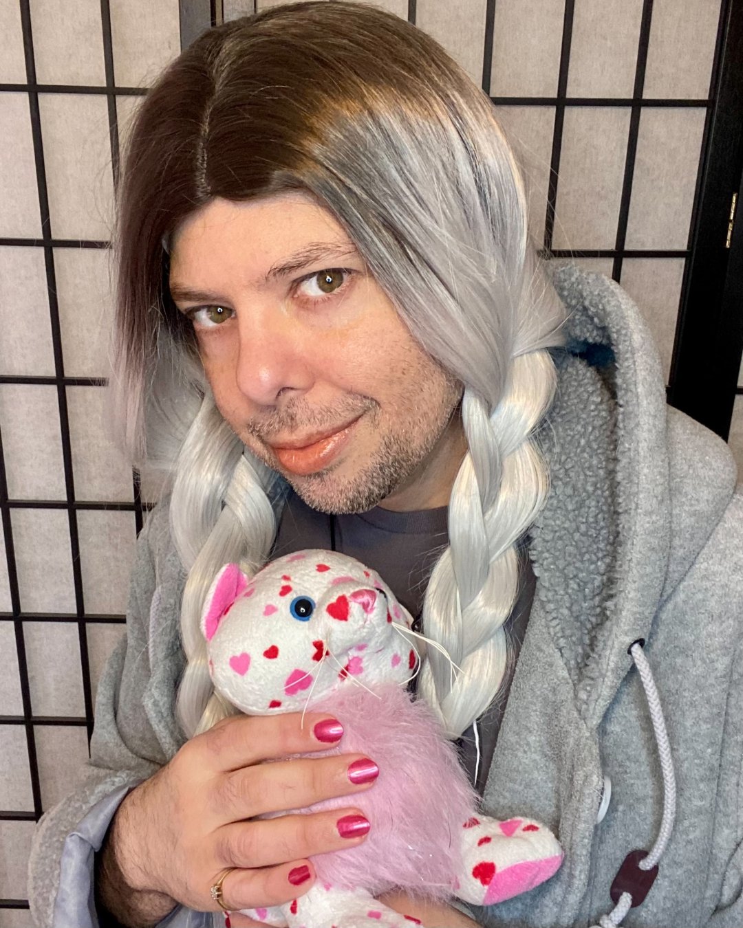 A person with long, braided hair in shades of brown and gray wears a gray coat. They are holding a stuffed animal with a pink and white design featuring hearts. Their nails are painted pink, and they have a subtle smile.